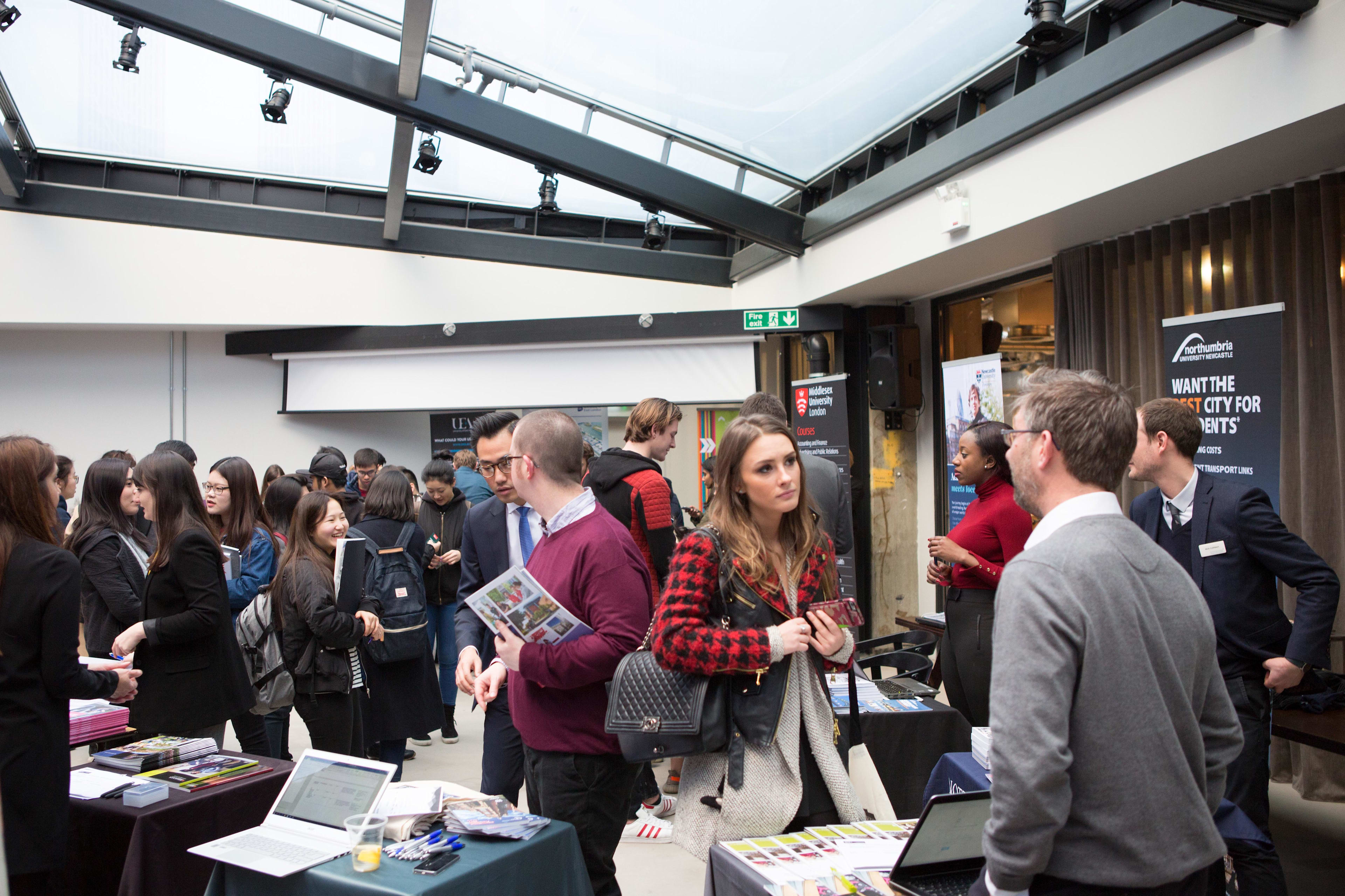 Image of room with University exhibition stands and international students talking to people about the universities