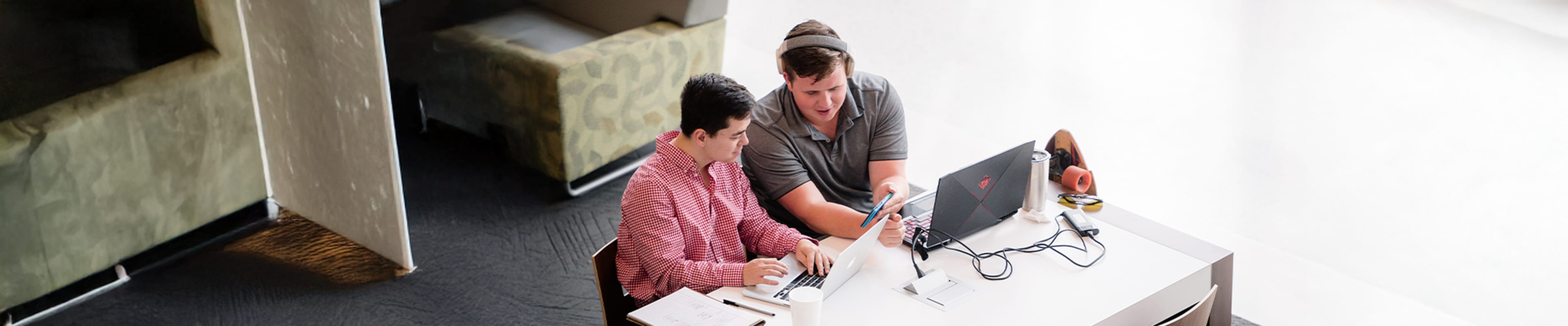 Two students working on their laptops while watching something in a mobile phone